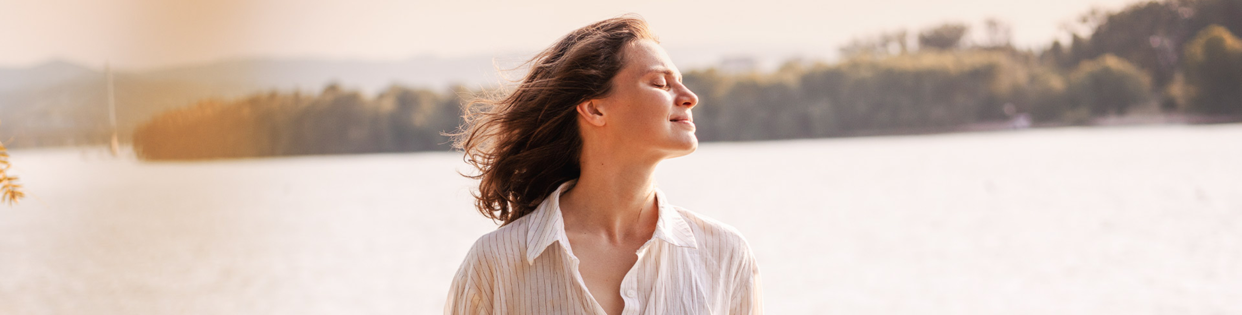 Žena stoji vani pored jezera i uživa u suncu zatvorenih očiju