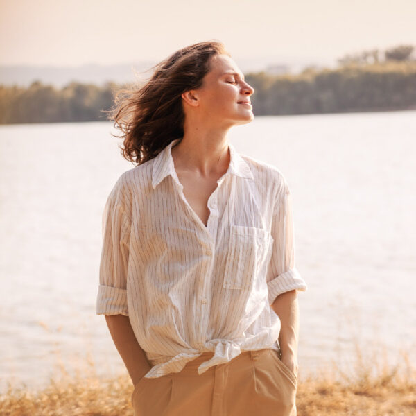 Žena stoji vani pored jezera i uživa u suncu zatvorenih očiju